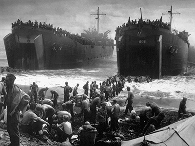 Soldiers disembark from large military ships onto a beach, with many personnel and equipment visible. The scene is bustling with activity, set against a cloudy sky.